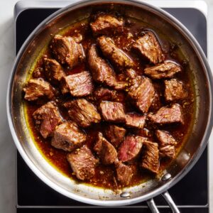 Top-down view of homemade Chinese beef searing in a stainless steel skillet on a black induction cooktop, with tender strips in glossy brown garlic sauce, some pieces still slightly pink, all glistening under natural light.