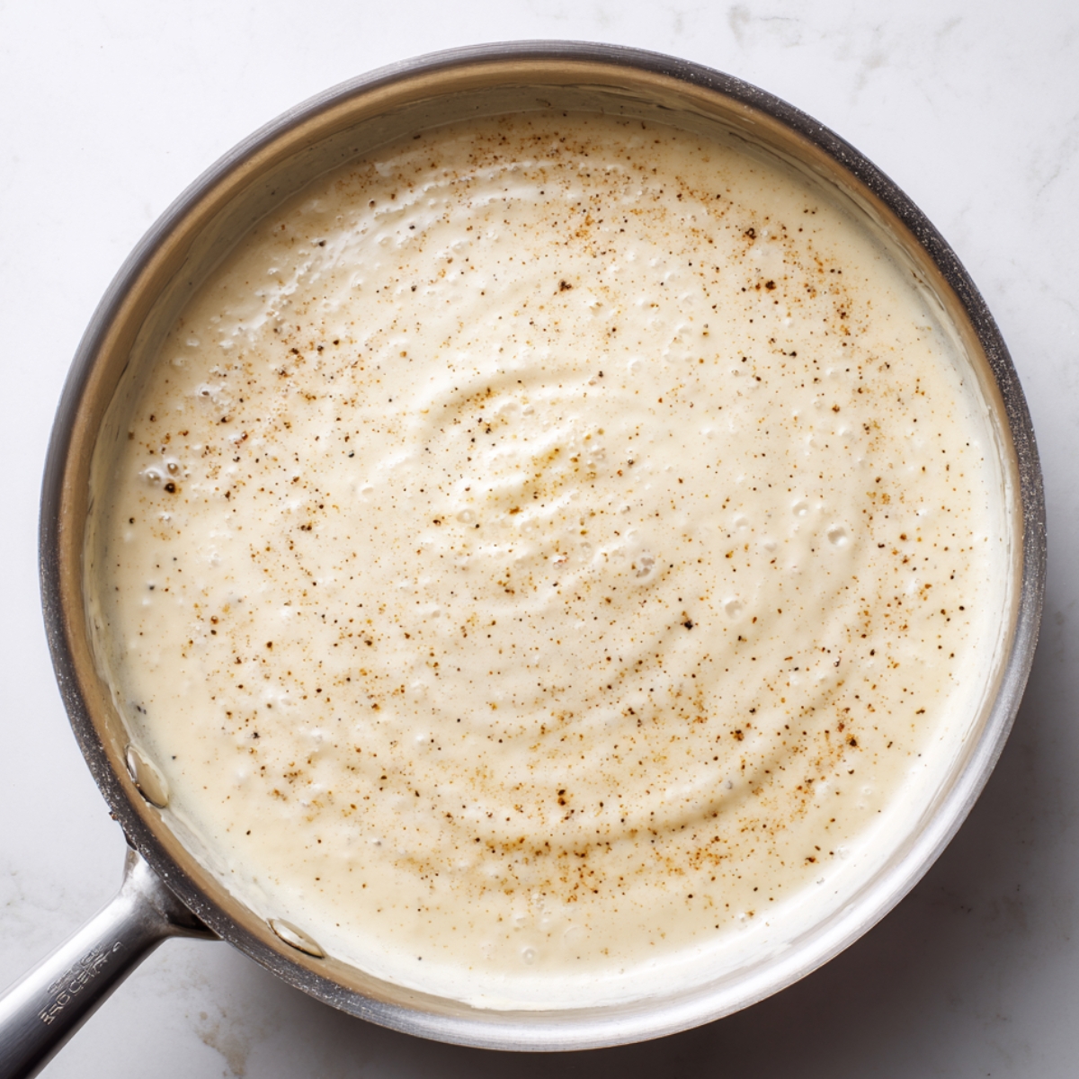 Stainless steel pan with creamy homemade alfredo sauce for seafood lasagna, sprinkled with black pepper and nutmeg on a white marble counter.