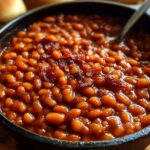 Golden, homemade baked beans in a round cast-iron Dutch oven, topped with crispy bacon and garnished with fresh thyme, showing a caramelized surface from slow oven baking.