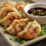 Homemade fried dumplings with crispy golden bottoms and pleated tops, garnished with green onions, served with soy dipping sauce in a white bowl.