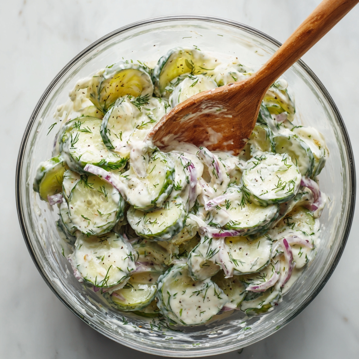 Homemade creamy cucumber salad with fresh dill and red onion in a glass bowl, stirred with a wooden spoon.
