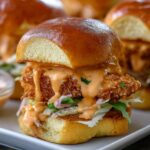Homemade Bang Bang Chicken Sliders on a white plate, with a glossy golden bun, crispy fried chicken, creamy orange bang bang sauce dripping down, fresh shredded lettuce, and herbs for garnish.