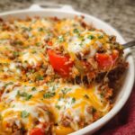 Homemade stuffed pepper casserole topped with melted cheddar and mozzarella, ground beef, rice, red and green bell peppers, and fresh herbs, with a wooden spoon lifting a cheesy serving.