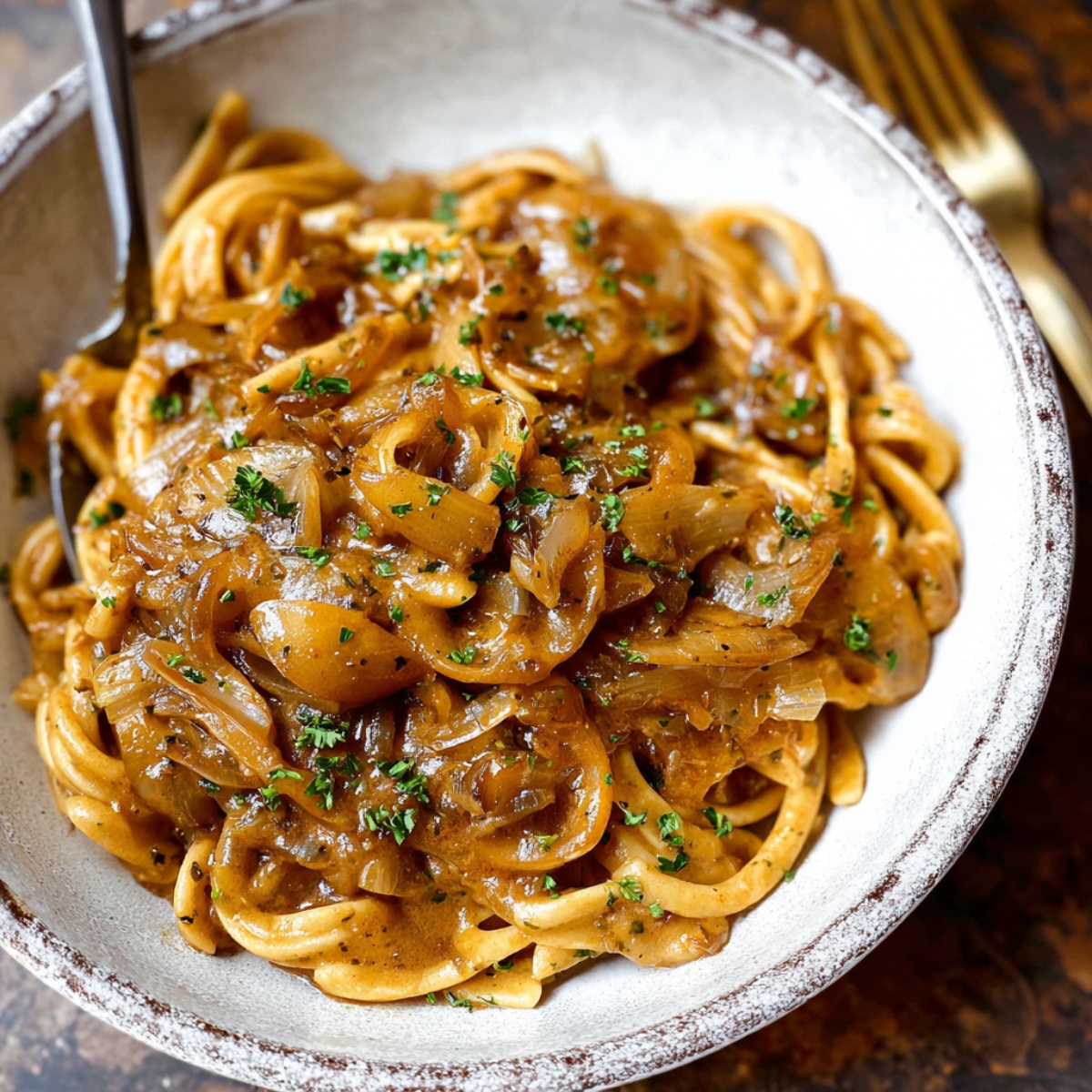 Bowl of homemade French onion pasta with caramelized onions and parsley in a rustic dish, golden fork in the background.