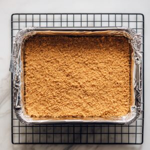 Unbaked graham cracker crust in a foil-lined pan on a cooling rack over a marble surface, smooth and golden, ready for baking.