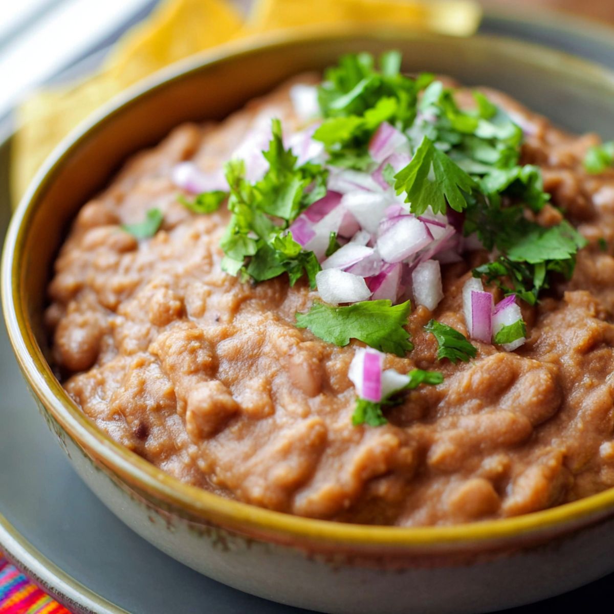 Bowl of homemade refried beans topped with fresh cilantro and diced red onion, served in a rustic ceramic dish on a gray plate with tortilla chips in the background.