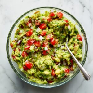 Chunky homemade guacamole with avocado, diced tomatoes, onion, cilantro, and black pepper in a glass bowl with a spoon, on a white marble countertop in natural light.