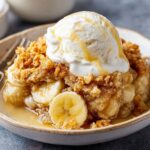 Banana Cobbler Recipe topped with vanilla ice cream and syrup, with visible banana slices and a golden crumb topping. A spoon rests in the bowl, and a whole banana is in the background.