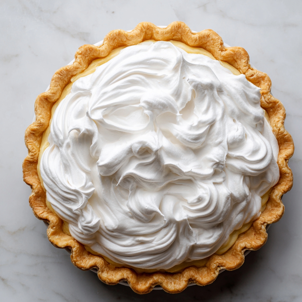 Homemade lemon meringue pie with a golden crust and glossy white meringue swirled into soft peaks on a marble countertop.