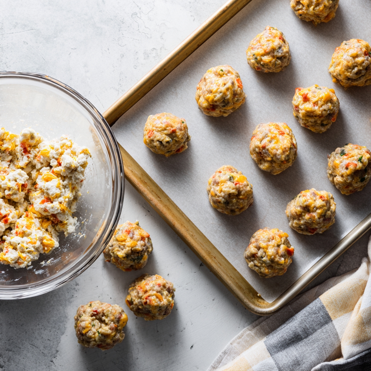 Unbaked Rotel Cream Cheese Sausage Balls on a parchment-lined baking sheet with a glass bowl of mixture and a plaid kitchen towel nearby.