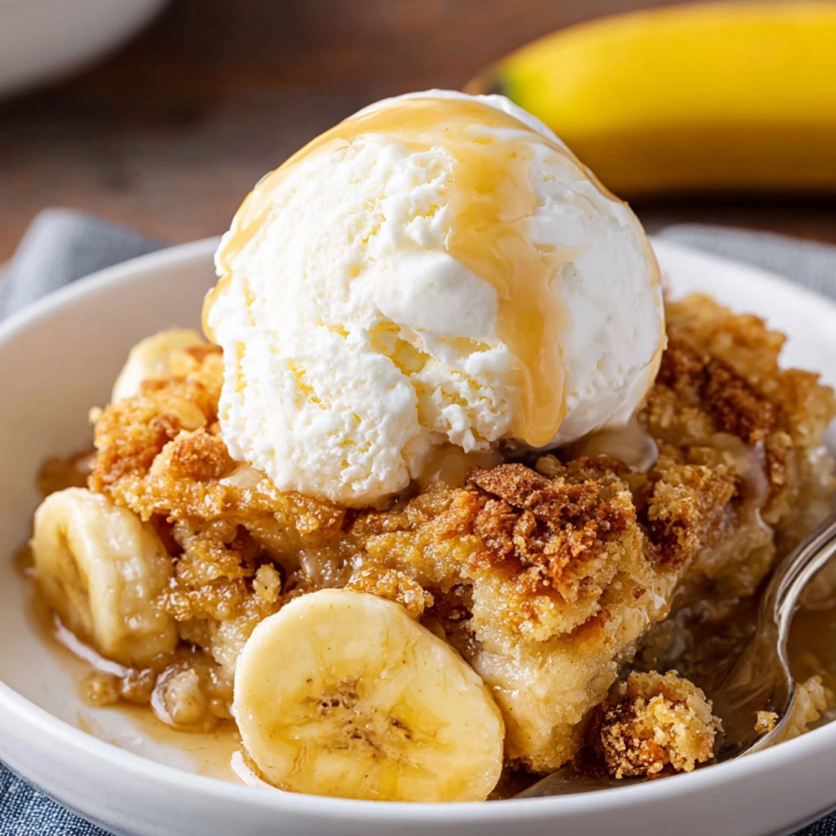 Homemade Banana Cobbler Recipe topped with vanilla ice cream and syrup, with visible banana slices and a golden crumb topping. A spoon rests in the bowl, and a whole banana is in the background.