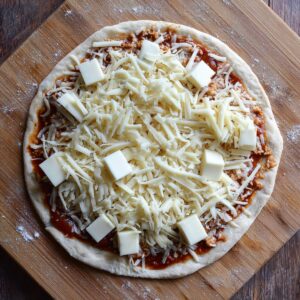 "Unbaked pizza with shredded and cubed mozzarella cheese on a layer of tomato sauce, on raw pizza dough placed on a wooden board."