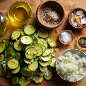 Chopped vegetables on a wooden board: green zucchini slices, yellow squash slices, thinly sliced red onions, and fresh herbs like parsley, with small bowls of spices.