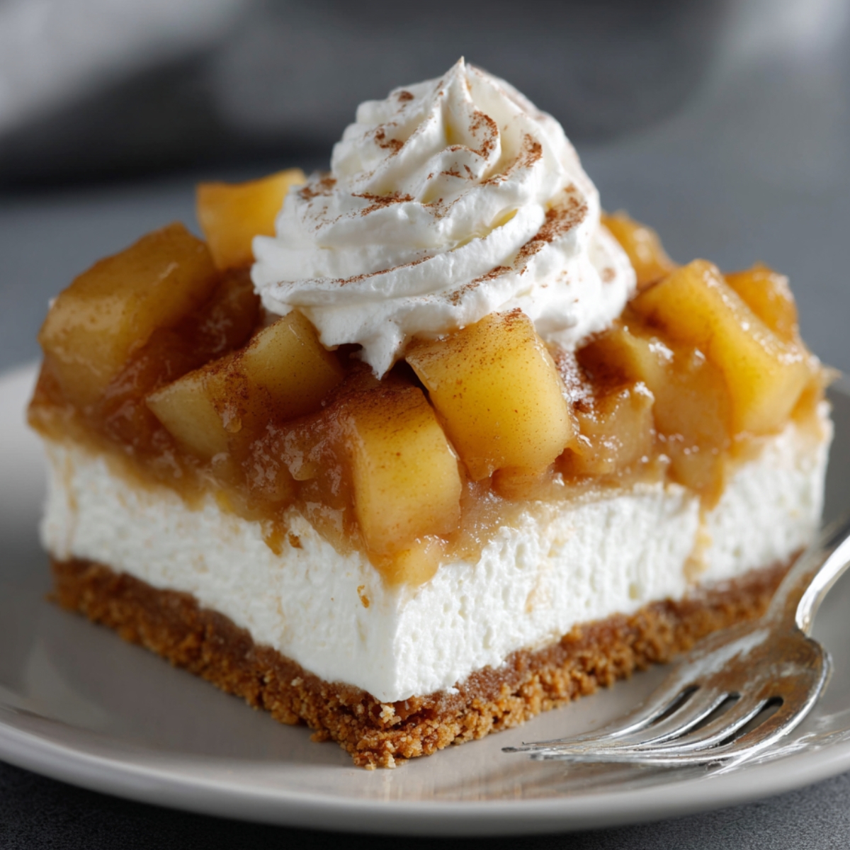 Caramel Apple Eclair Cake with a graham cracker crust, creamy filling, caramelized apples, and whipped cream topped with cinnamon, served on a beige plate with a blurred red apple in the background.