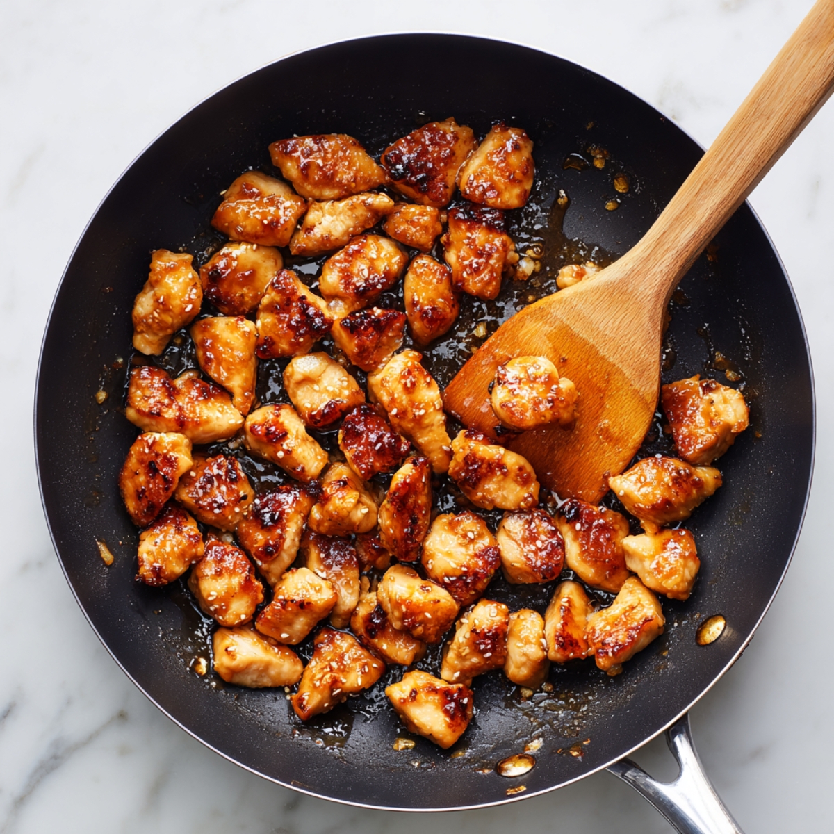 Golden-brown teriyaki chicken pieces cooking in a black skillet with a wooden spatula on a marble counter, glossy and homemade.