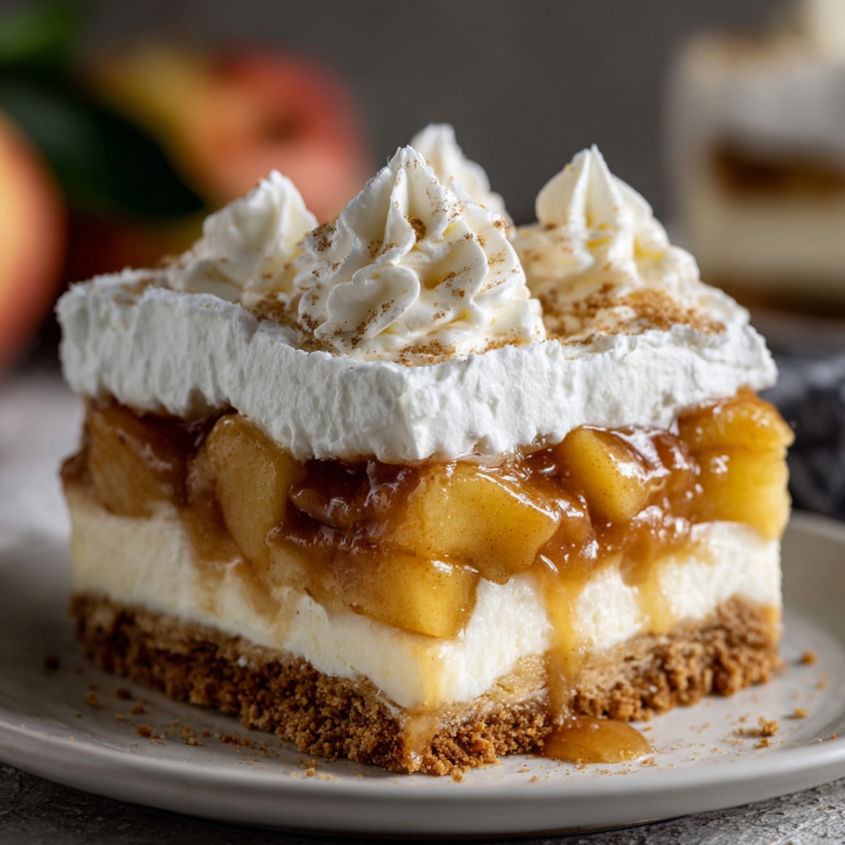 A slice of homemade Caramel Apple Eclair Cake with a graham cracker crust, creamy filling, caramelized apples, and whipped cream topped with cinnamon, served on a beige plate with a blurred red apple in the background.