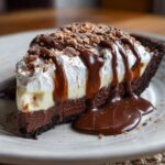 Mississippi Mud Pie slice with chocolate cookie crust, fudgy chocolate layer, pudding, and whipped cream, topped with chocolate drizzle and shavings on a white plate.