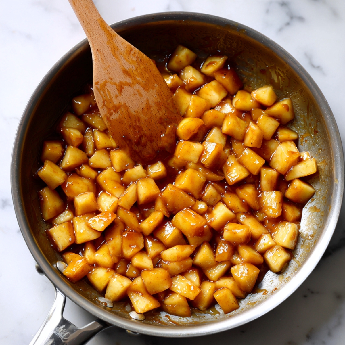 A skillet of caramelized diced apples in syrup, stirred with a wooden spoon on a marble surface.