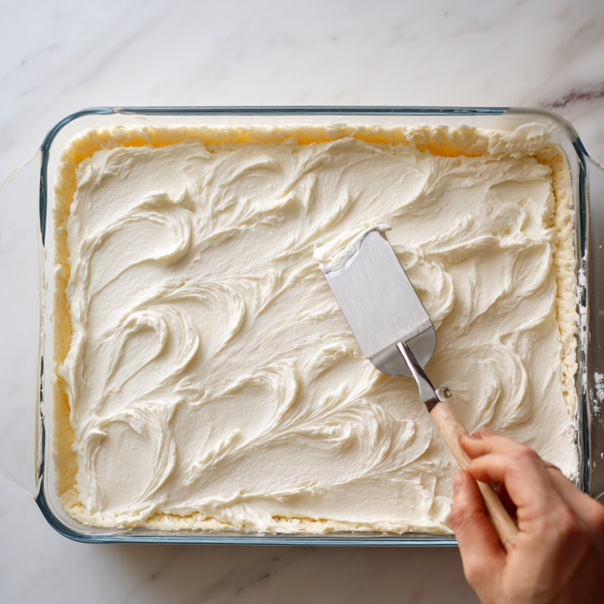 A hand uses an offset spatula to spread creamy cheesecake filling evenly over a layer of dough in a glass baking dish, creating soft swirls on the smooth surface, on a bright marble countertop.