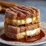 Homemade Churro Cheesecake dessert stacked with two layers of cinnamon-sugar churro cake filled with creamy cheesecake and drizzled generously with caramel sauce on a plate.