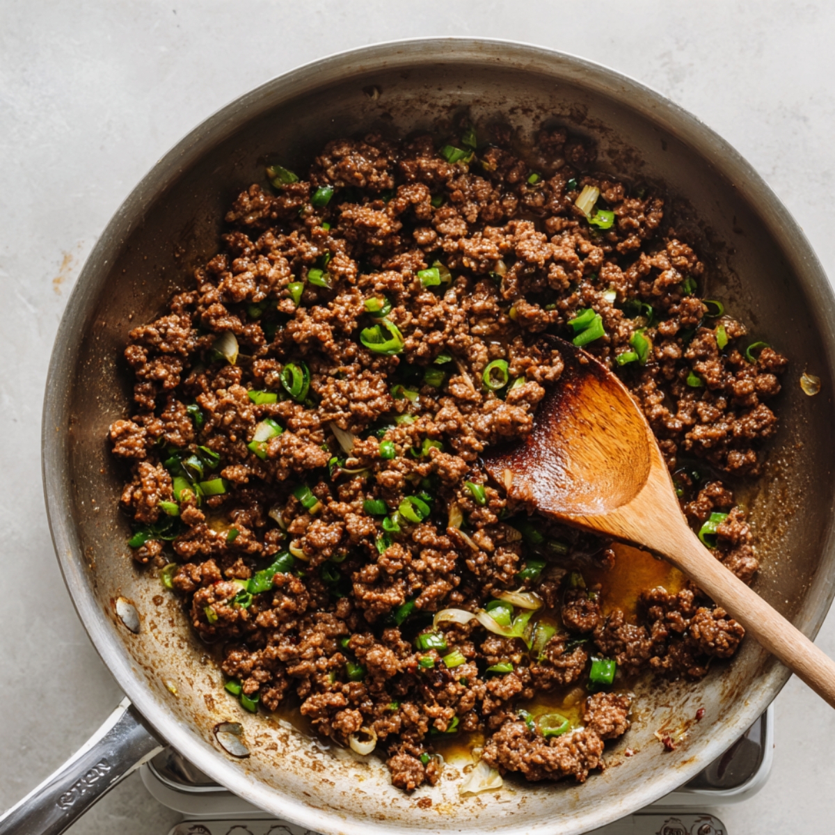 Homemade Korean ground beef cooking in a skillet with chopped green onions and sauce, stirred with a wooden spoon.
