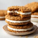 Homemade Oatmeal Cream Pies with one showing a bite taken out, revealing the creamy filling between the oatmeal cookies. The cookies are golden brown and placed on a simple ceramic plate.