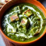 A bowl of creamy Palak Paneer with paneer cubes in a vibrant spinach gravy, topped with a swirl of cream, served with naan.