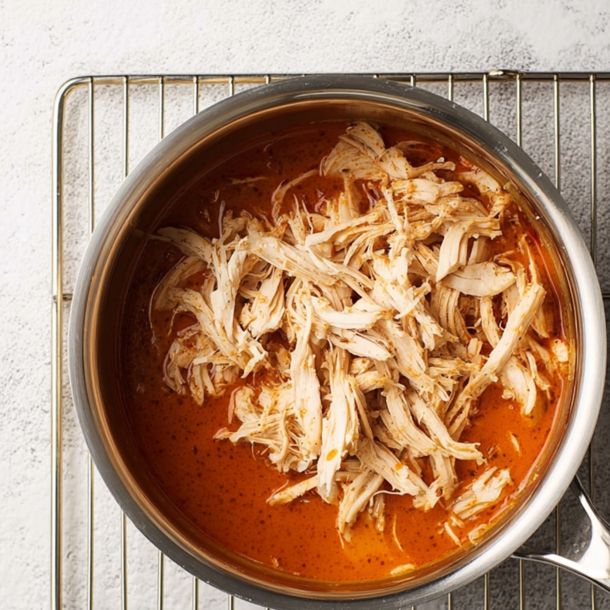 Overhead view of shredded chicken being added into a pan of orange buffalo sauce, beginning to mix into the sauce on a wire rack over a light kitchen countertop.