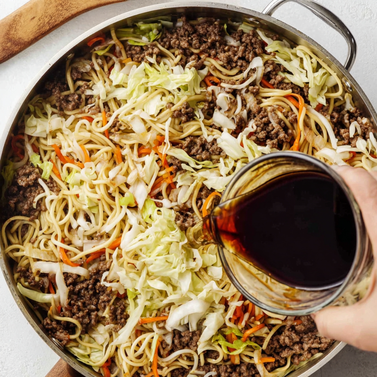 Hand pouring dark stir-fry sauce into a skillet with noodles, ground pork, cabbage, and carrots before mixing