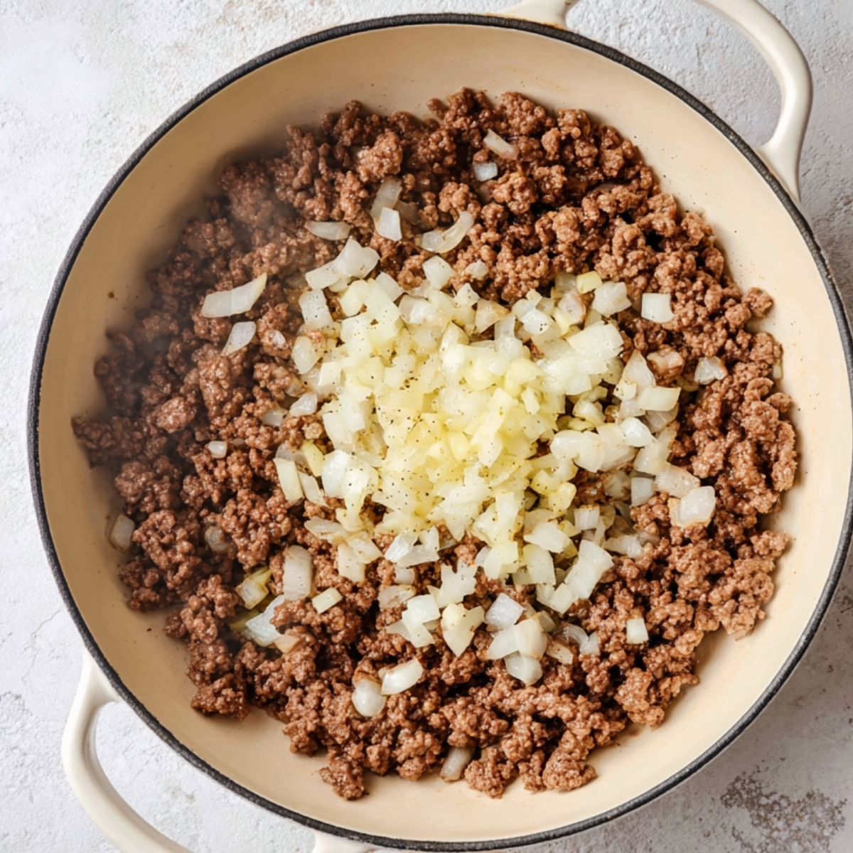 Browned ground beef in a skillet topped with diced onion and garlic before mixing, overhead cooking step
