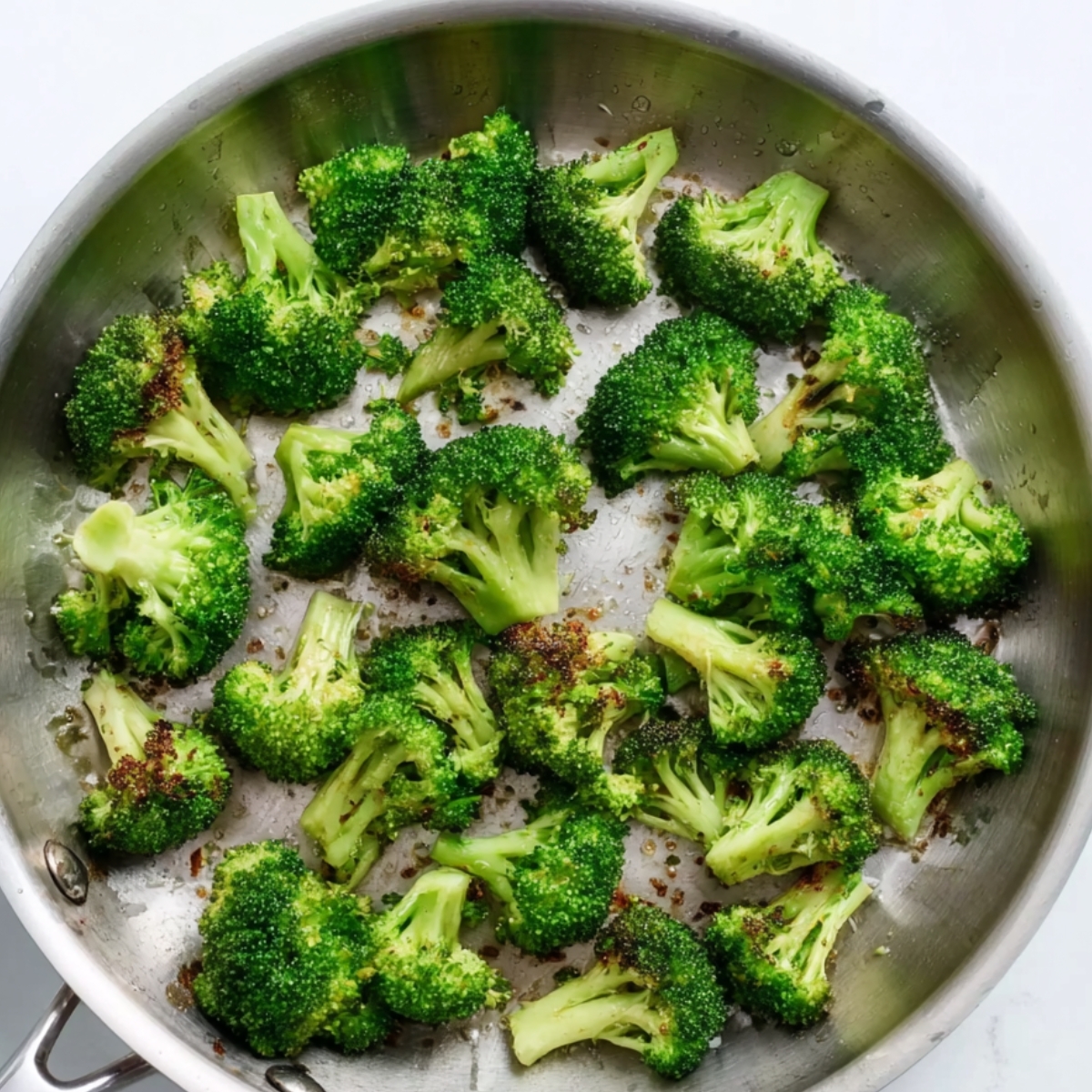 Fresh broccoli florets sautéing in a stainless steel skillet until lightly browned and tender, photographed from above on a clean white background.