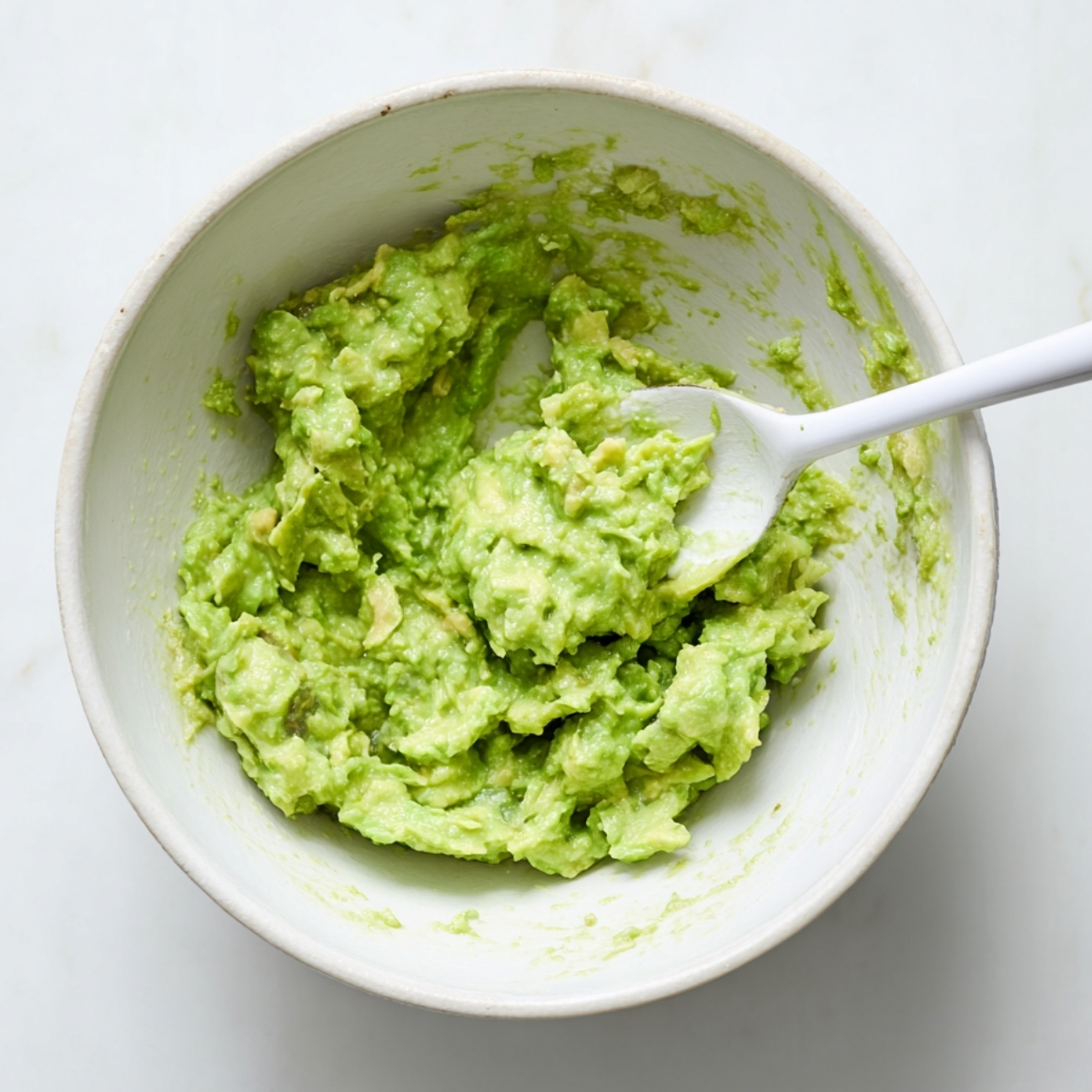 Bowl of freshly mashed guacamole with visible avocado chunks and a spoon, shot overhead on a white background.