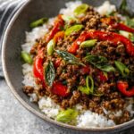 A completed dish of Thai basil beef served over white rice in a gray bowl. The dish features ground beef, red bell peppers, and fresh basil leaves, garnished with sliced green onions.