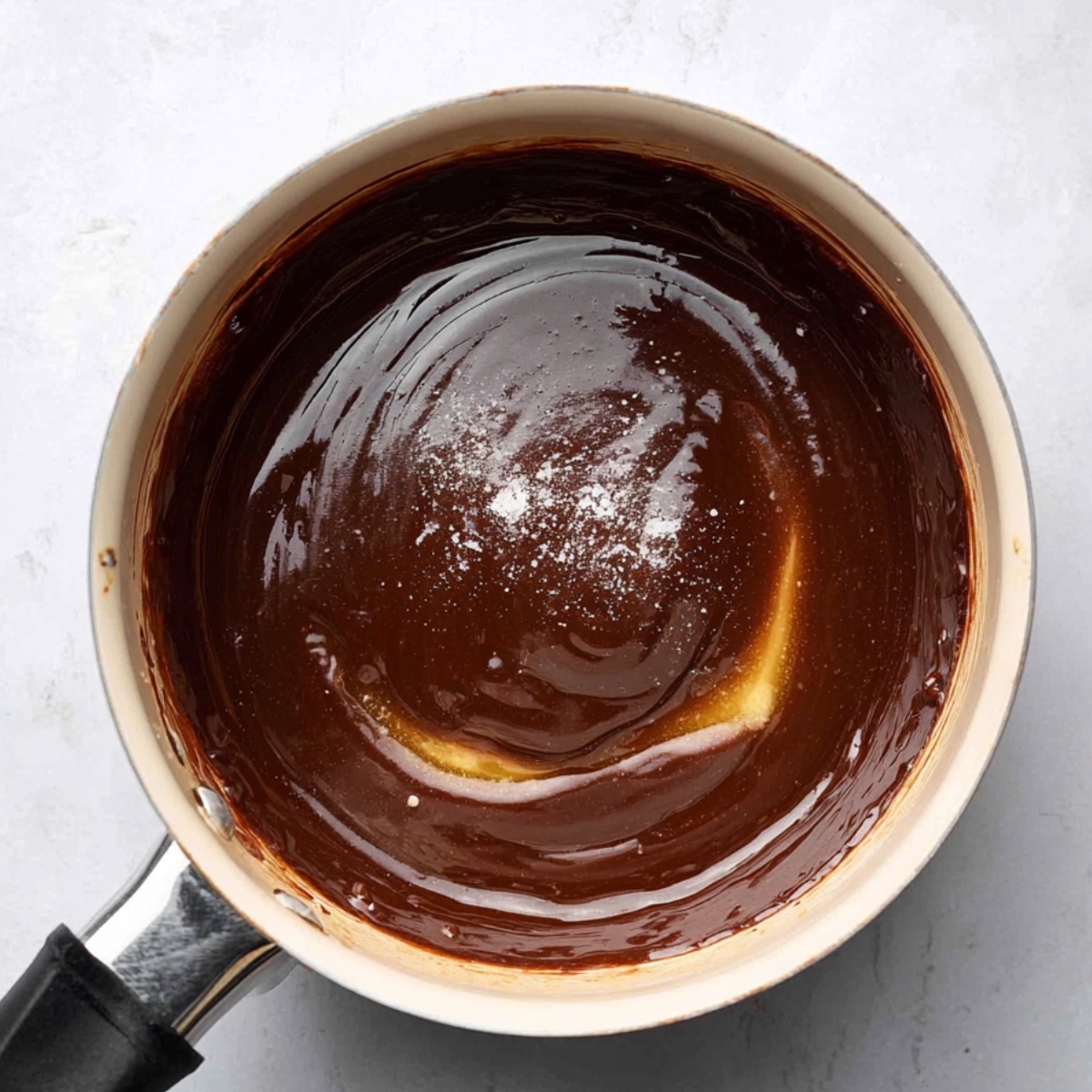 A saucepan with a glossy, thick chocolate mixture, showing added vanilla extract and a pinch of salt.