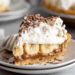 A close-up of a decadent slice of Banoffee Pie, topped with whipped cream, chocolate shavings, and a layer of banana slices on a buttery graham cracker crust.