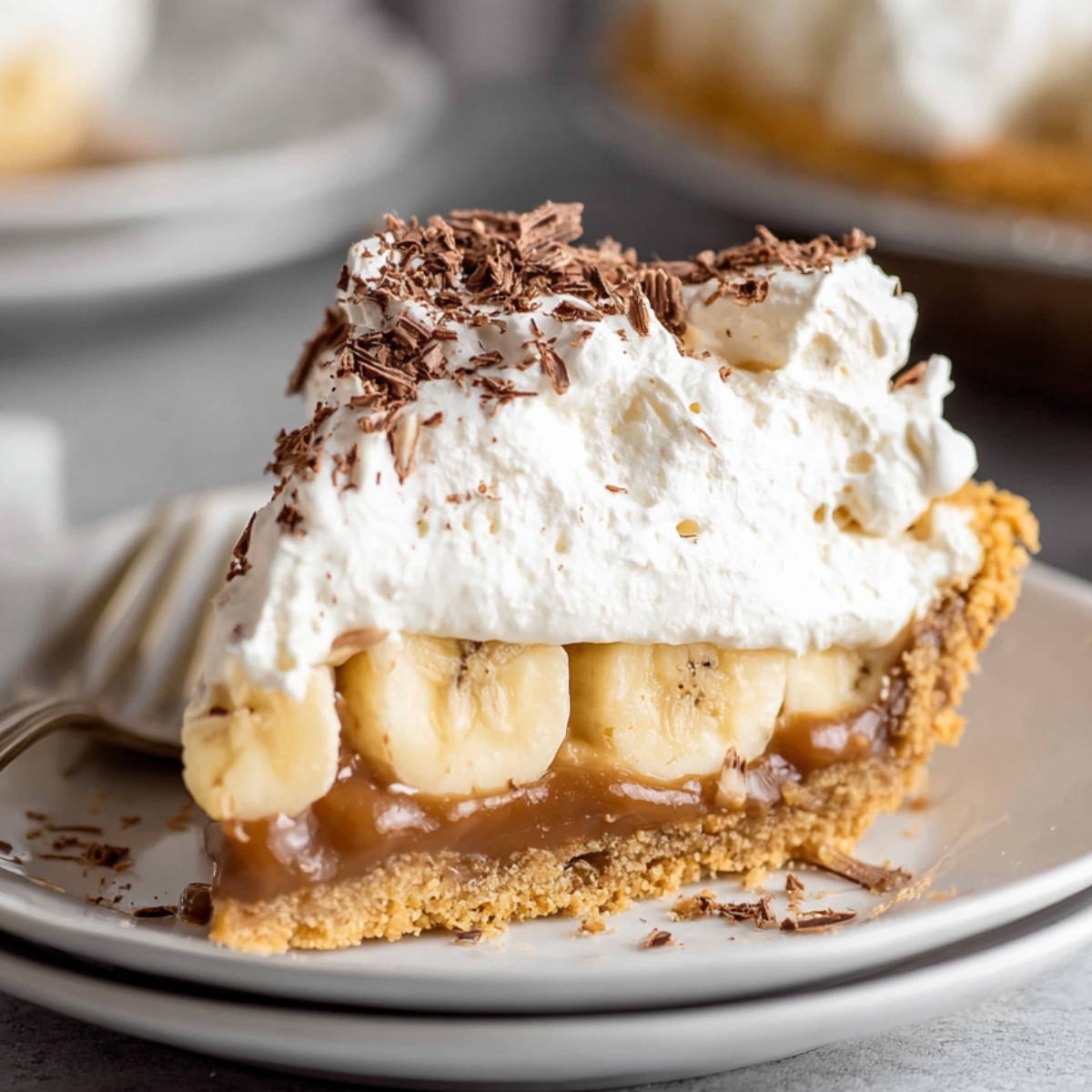 A close-up of a decadent slice of Banoffee Pie, topped with whipped cream, chocolate shavings, and a layer of banana slices on a buttery graham cracker crust.