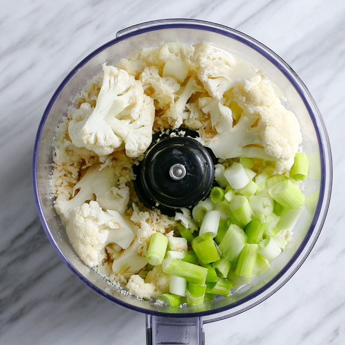 A close-up of cauliflower florets and green onions in a food processor, prepared for pulsing into a rice-like texture for Cauliflower Tabbouleh.