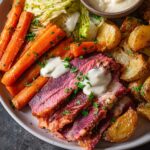 A plate of sliced Corned Beef and Cabbage served with roasted potatoes, glazed carrots, and sautéed cabbage, topped with a creamy sauce and garnished with parsley.