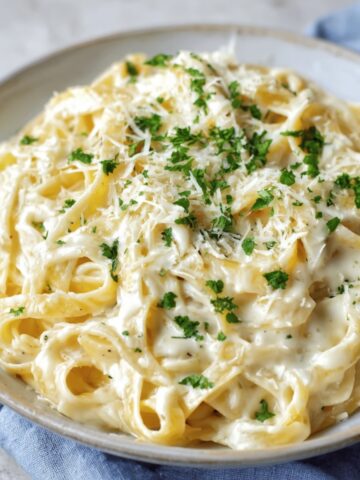Plate of creamy Fettuccine Alfredo topped with fresh parsley and grated parmesan cheese. The pasta is rich and creamy, served on a light blue napkin.