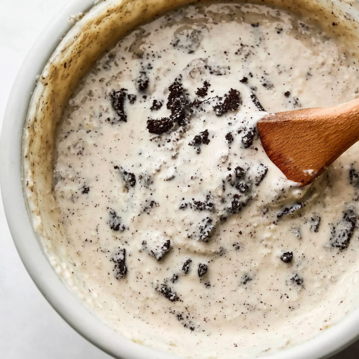 A bowl of cake batter mixed with chunks of Oreo cookies, with a wooden spatula for stirring.