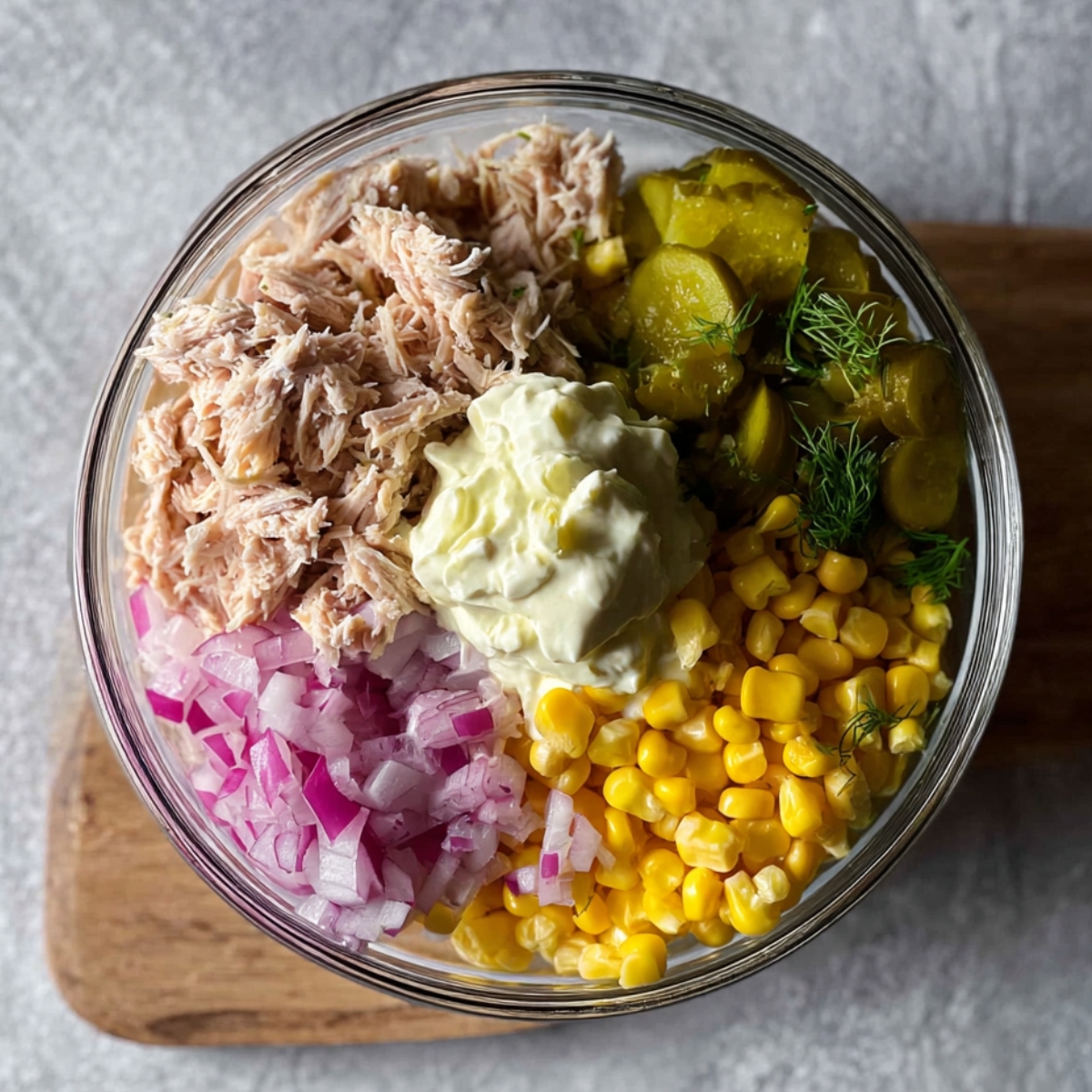 A glass bowl filled with ingredients for a tuna filling, including shredded tuna, corn, pickles, onions, and mayonnaise.