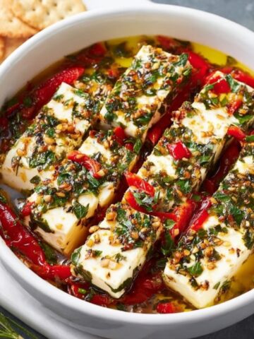 A dish of Marinated Cheese, including slices of cheese and red peppers soaked in an aromatic herb oil marinade.