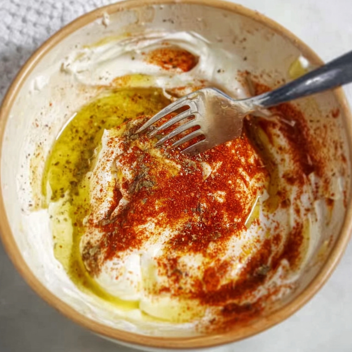 A bowl filled with yogurt, olive oil, and spices like paprika, being mixed with a fork to create a flavorful dipping sauce for the kofta kebabs.