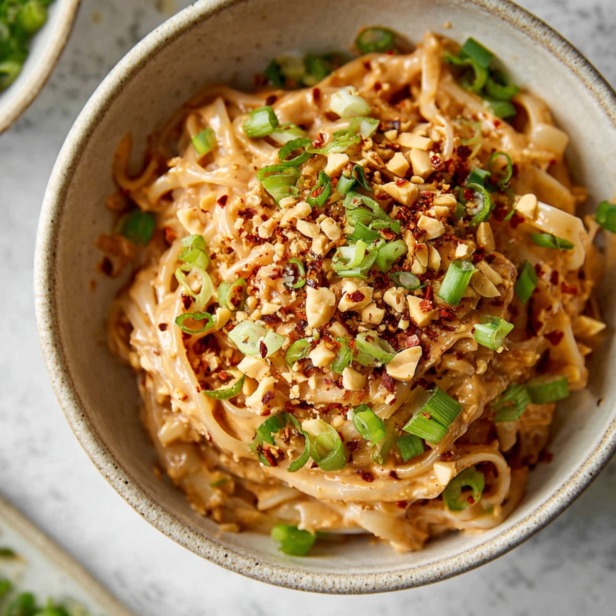 A bowl of Spicy Peanut Butter Noodles topped with green onions, crushed peanuts, and red chili flakes, creating a vibrant and flavorful dish.