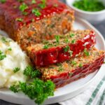 A plate of Vegan Lentil Loaf slices, with mashed potatoes and green peas, topped with parsley and glazed with ketchup.