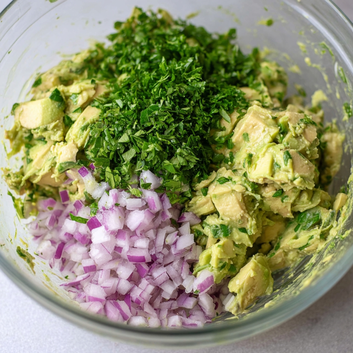 A glass bowl containing mashed avocado, finely chopped red onion, and fresh cilantro, ready to be mixed into a flavorful filling for wraps.