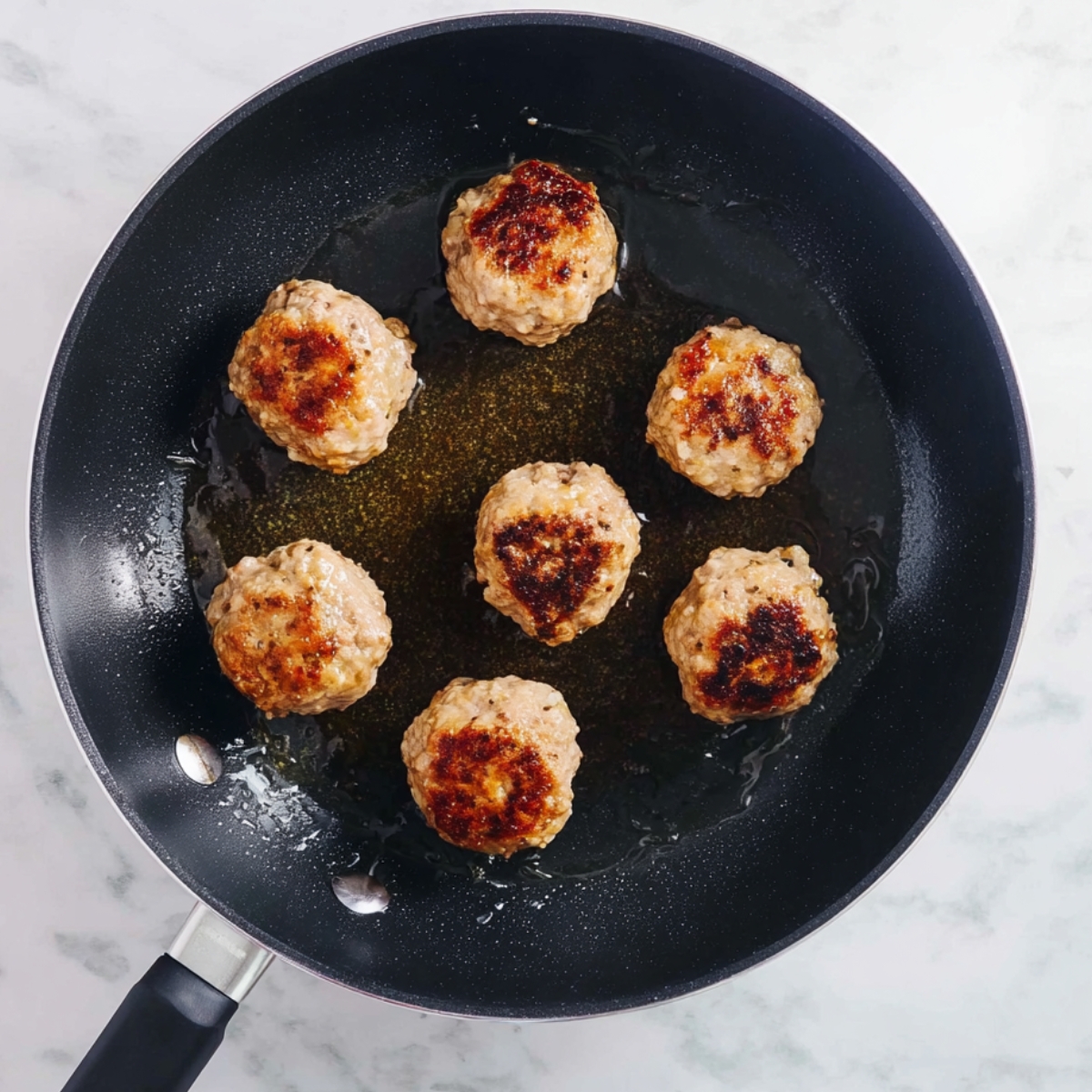 A skillet filled with golden-brown meatballs, lightly browned on one side, cooking in a layer of oil, with a neutral marble background.