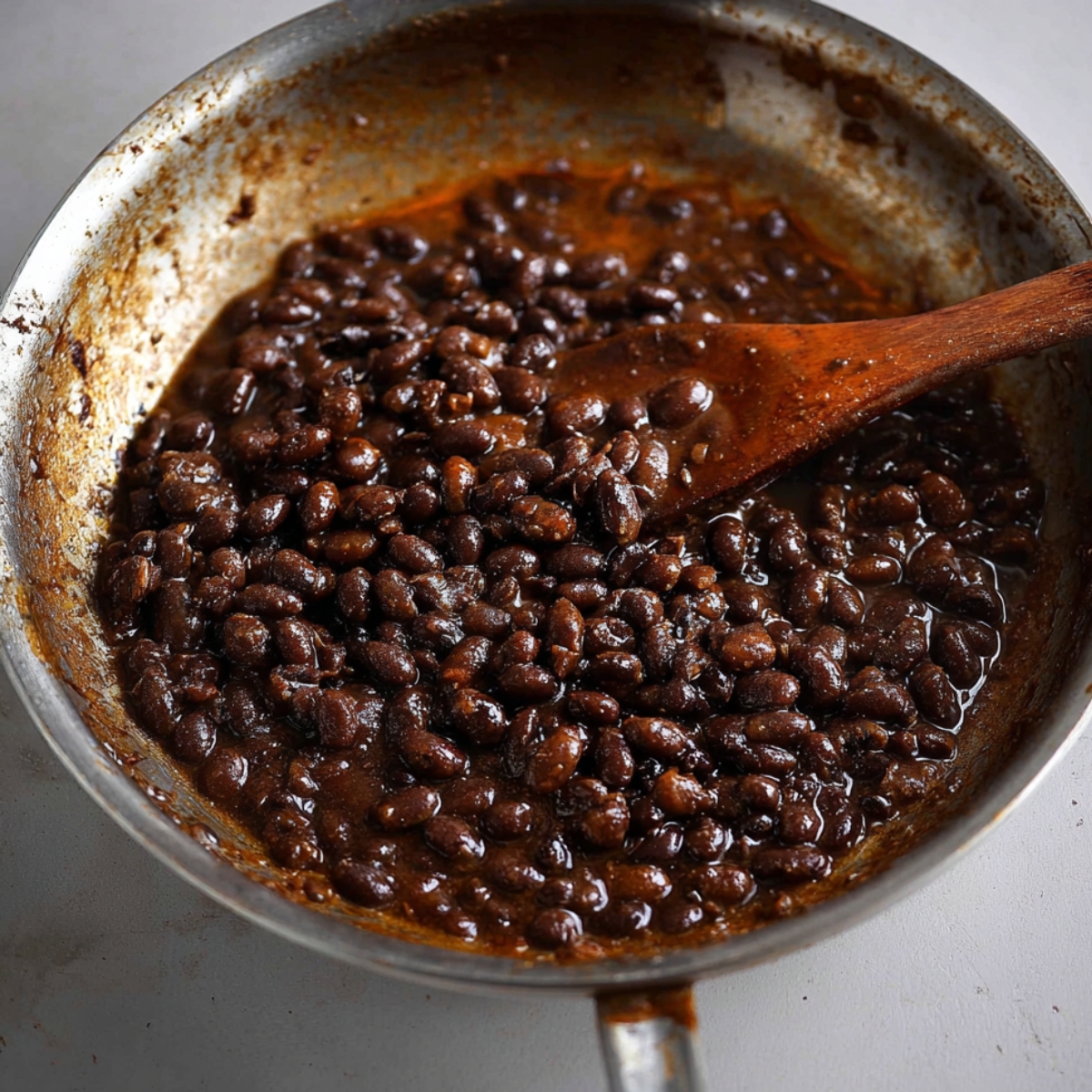 A pan with black beans cooked in a sauce, with a wooden spoon stirring the mixture.