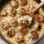 A close-up of Creamy Marry Me Chicken Meatballs in a skillet, simmering in a rich sauce, with a wooden spoon lifting one meatball, surrounded by a smooth, savory sauce.