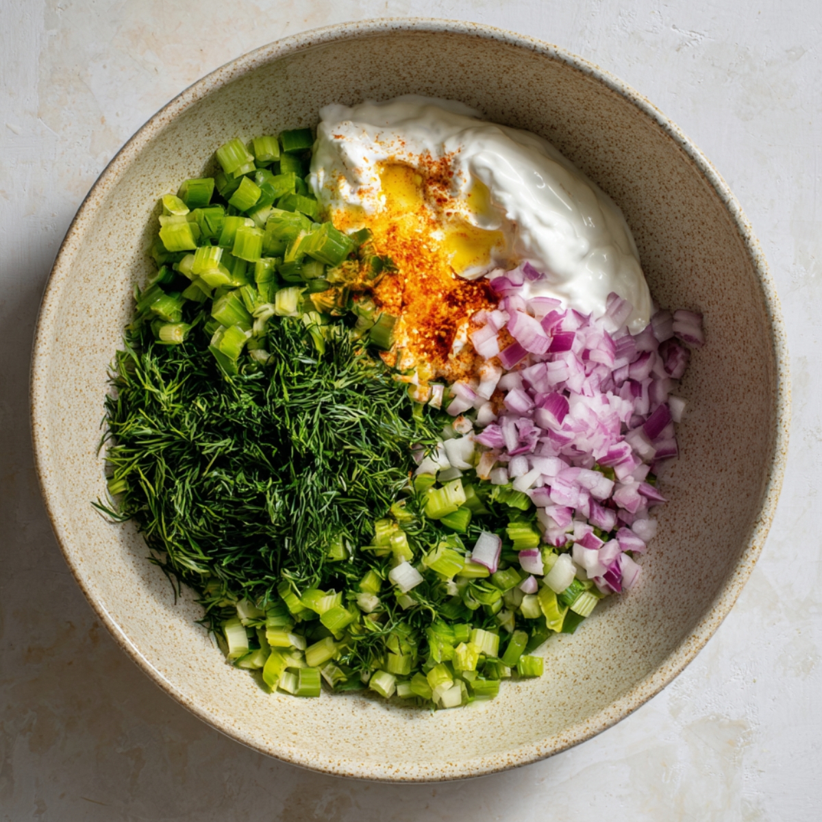 Ingredients for a sauce including yogurt, diced celery, red onions, and spices, mixed together in a bowl.